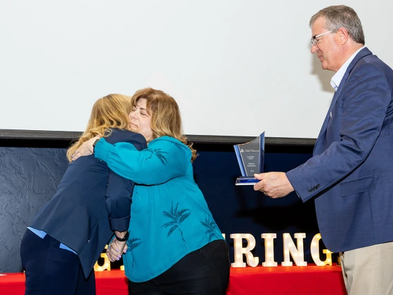 Two women in blue shirts embrace for a hug and one man in a blue blazer holds an award.