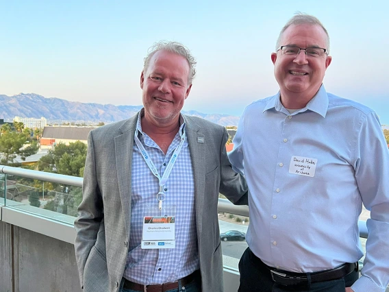 Two men with gray hair and blue dress shirts pose for a photo in front of a mountain range.