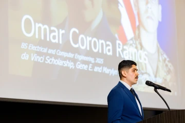 A brown haired man stands in front of a screen with his picture and name depicted.