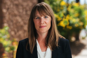 A woman with brown hair and curtain bangs poses for a photo in front of greenery.