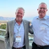 Two men with gray hair and blue dress shirts pose for a photo in front of a mountain range.